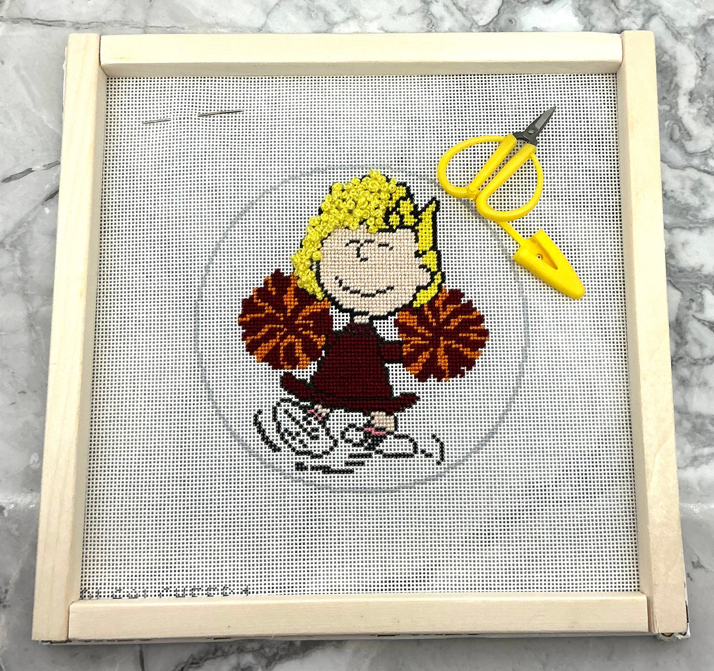 Needlepoint design of Sally from Peanuts as a cheerleader with a pair of yellow scissors